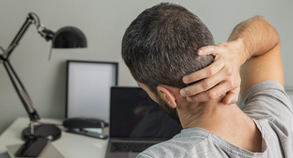 Hombre sentado en un escritorio con postura encorvada, sujetándose la parte baja de la espalda y el cuello, mostrando signos de dolor muscular. Convendría realizar ejercicios para evitar lesiones en la vuelta al trabajo.