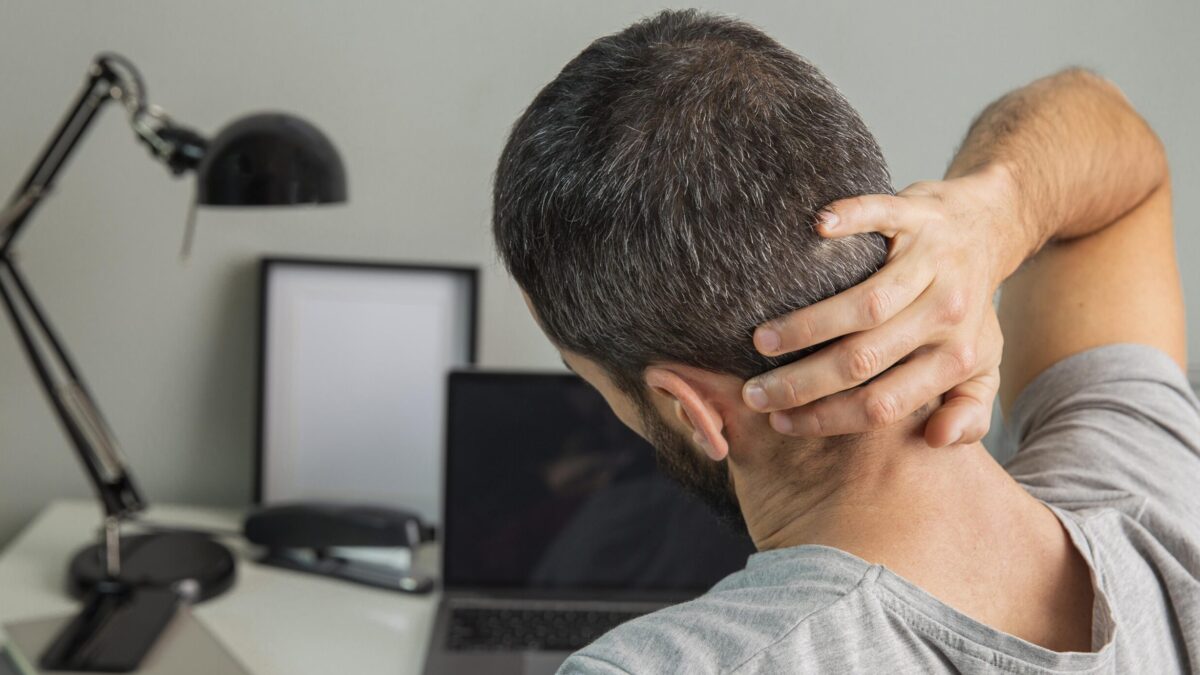 Hombre sentado en un escritorio con postura encorvada, sujetándose la parte baja de la espalda y el cuello, mostrando signos de dolor muscular. Convendría realizar ejercicios para evitar lesiones en la vuelta al trabajo.
