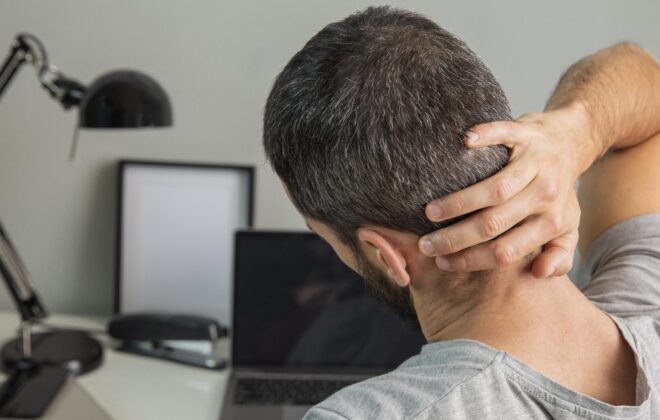 Hombre sentado en un escritorio con postura encorvada, sujetándose la parte baja de la espalda y el cuello, mostrando signos de dolor muscular. Convendría realizar ejercicios para evitar lesiones en la vuelta al trabajo.