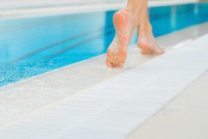 Imagen de unos pies descalzos en el borde de una piscina. Este entorno, habitual en verano, puede causar problemas podológicos como durezas, contracturas o verrugas plantares por el contacto prolongado con superficies duras, húmedas o calientes. Representa una de las situaciones más frecuentes que tratamos en Centro Fisher.